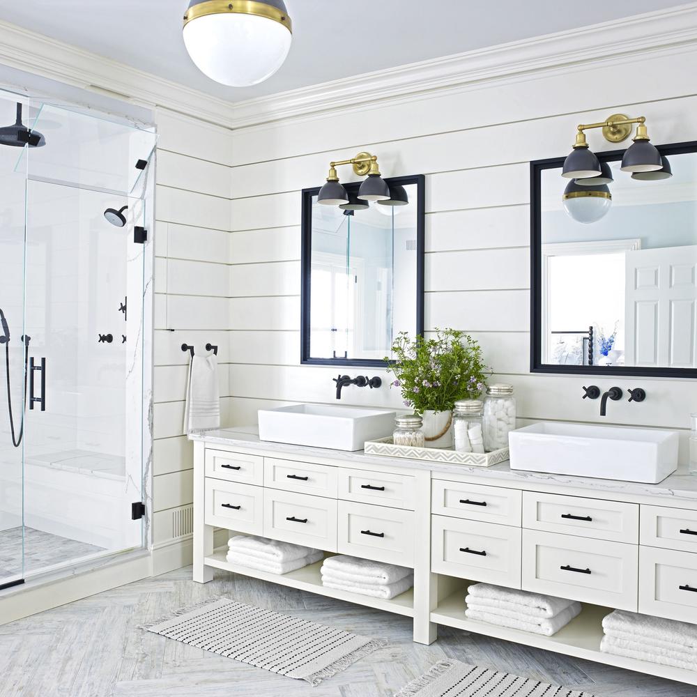 A spacious white bathroom featuring a dual sink vanity with black handles and mirrors, complemented by unique brass fixtures and cozy accessories.