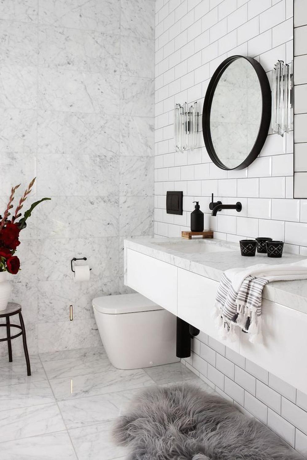 A modern bathroom design featuring black fixtures and white marble tiles. It includes a floating vanity and round black-framed mirror.