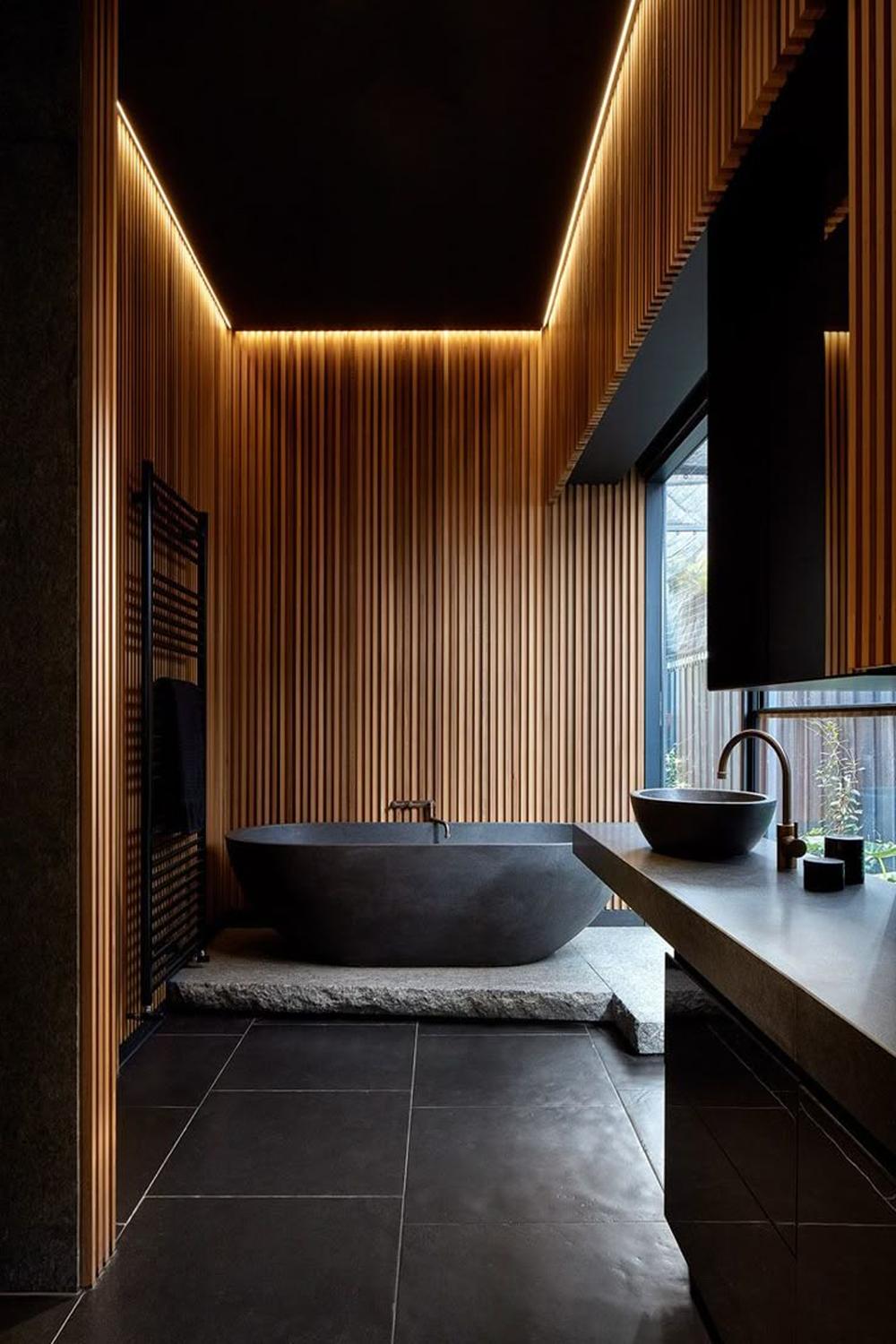 Sophisticated bathroom featuring a freestanding black stone tub, vertical wooden wall accents, and warm LED lighting.