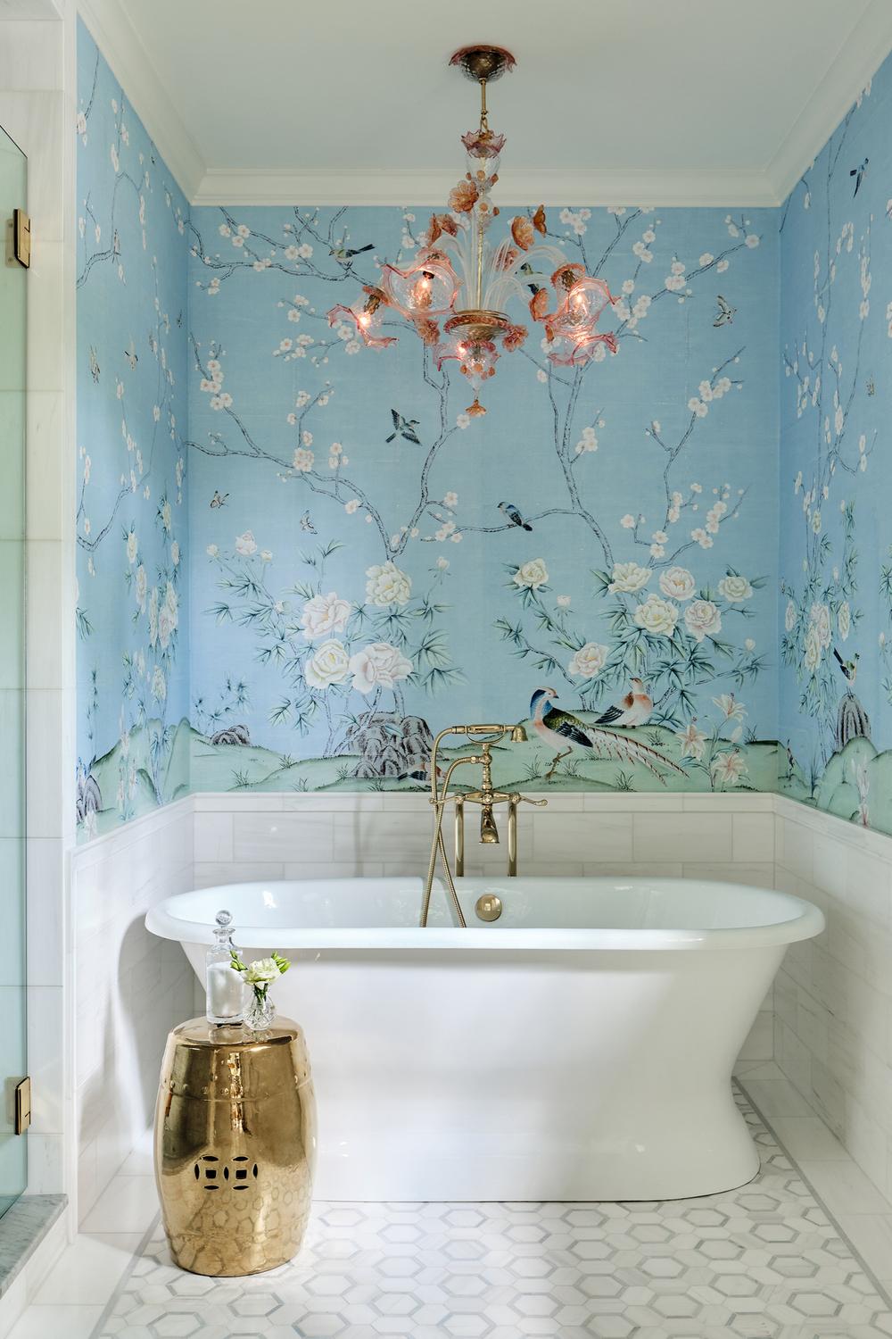 Light blue Chinoiserie wallpaper adorns a bathroom featuring a modern freestanding tub and a stylish chandelier, delivering calm elegance.
