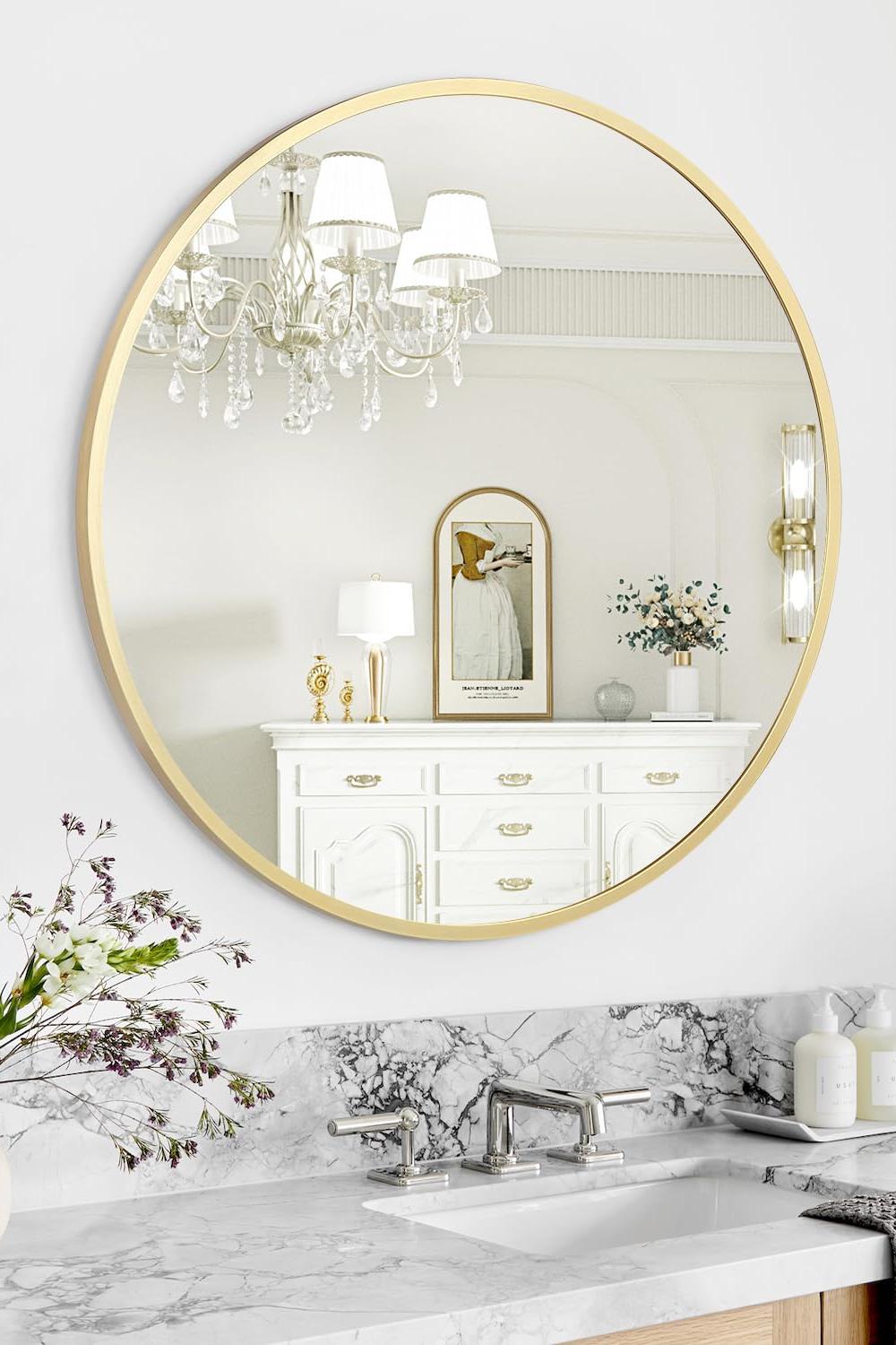 Elegant gold framed round bathroom mirror above a marble countertop with a chic dresser and floral decor.