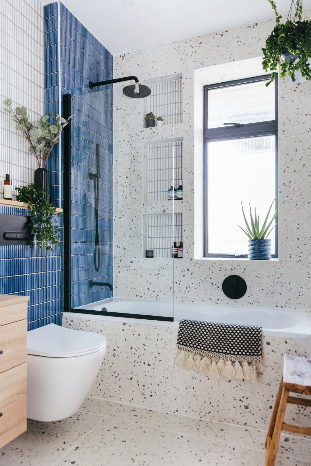 Stylish modern small bathroom featuring a bathtub integrated with a shower enclosure, bold blue and speckled terrazzo wall tiles, and natural light from a window.