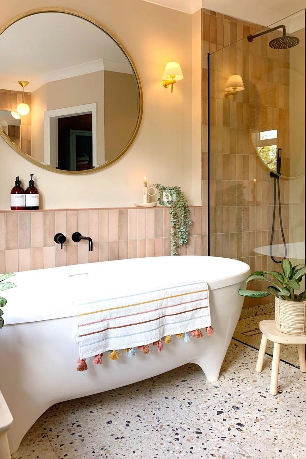 A warm bathroom featuring soft pastel tones, a freestanding bathtub, and a round mirror framed in gold.