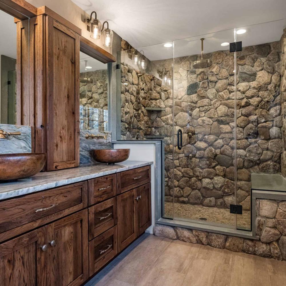 Sophisticated stone-themed bathroom with a rustic stone wall, spacious glass-enclosed shower, double vanity with warm wood finish, and round bowl sinks.