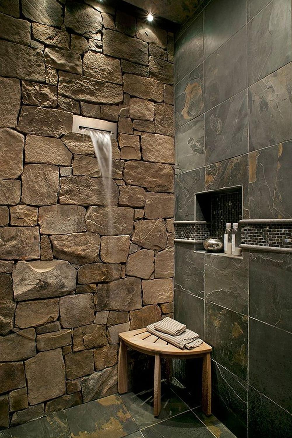 Elegant stone bathroom showcasing textured stone wall with integrated water feature, spacious shower area with recessed lighting, and rustic wooden bench.