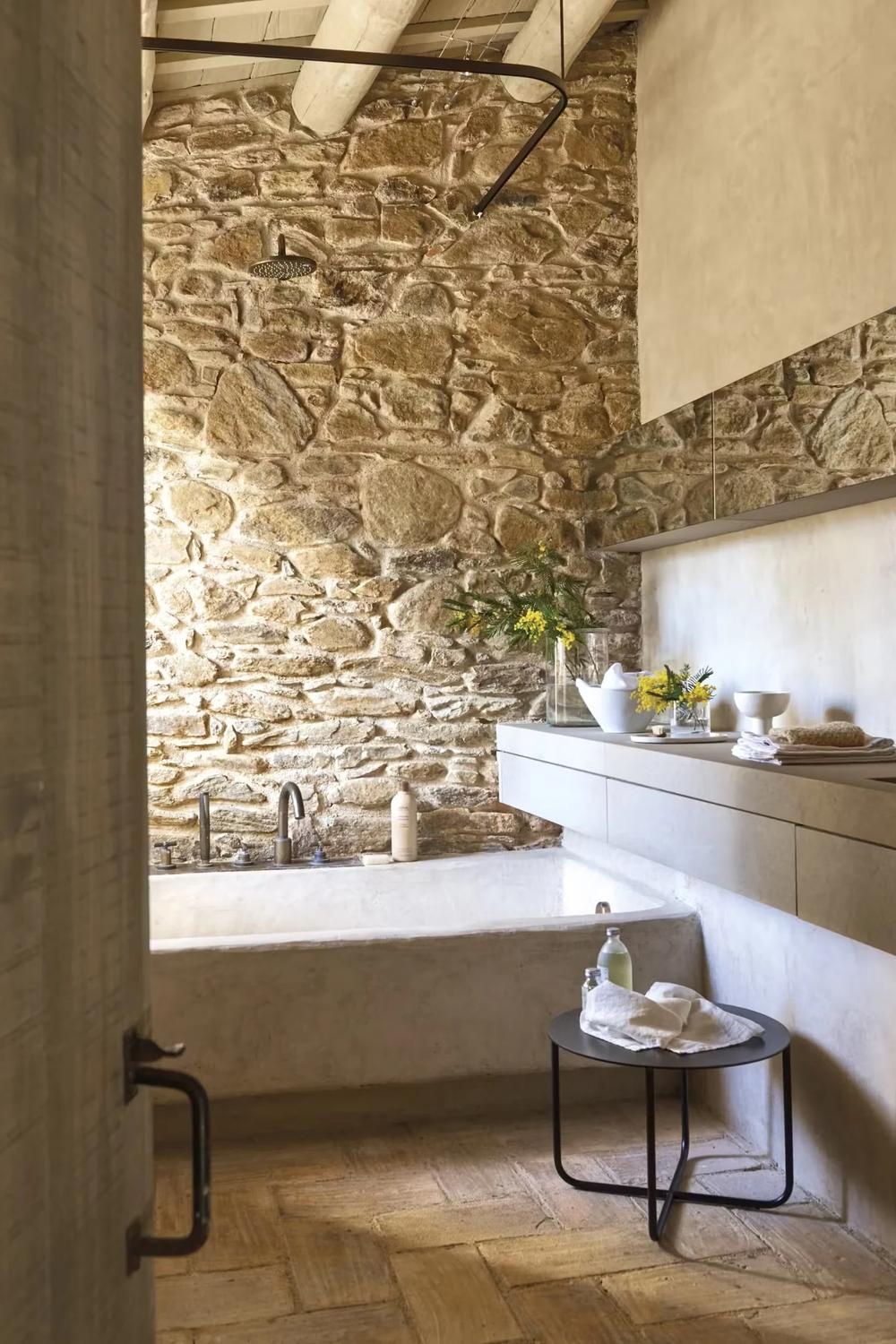 Bathroom boasting freestanding white bathtub, rustic stone wall, and modern rain showerhead.