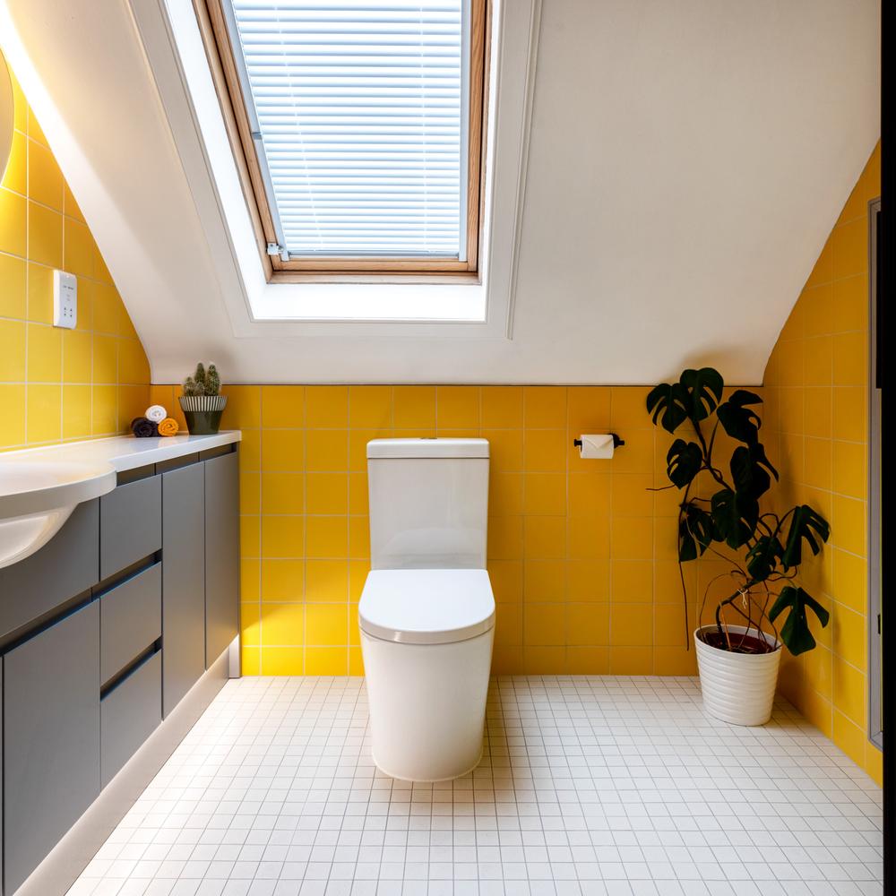 A modern bathroom featuring vibrant yellow tiles, roof window, gray vanity, decorated with towels and plants for a cheerful atmosphere.
