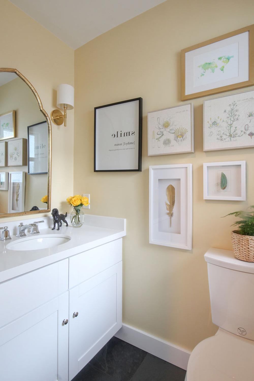 A cozy bathroom with soft yellow walls, white vanity, golden accents, and artful decor elements, creating a warm atmosphere.